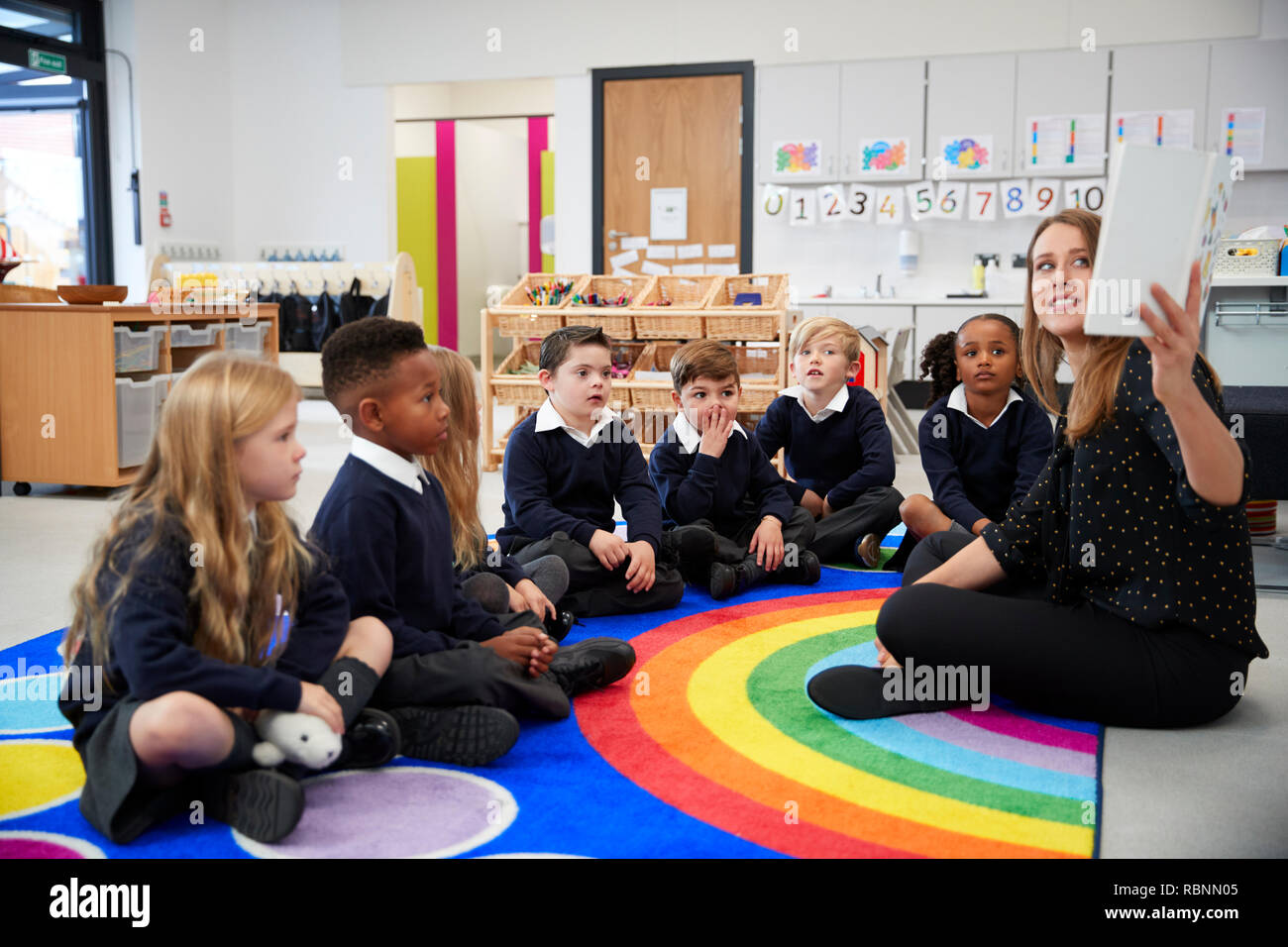 Lehrerin hält ein Buch vor ihrer Klasse der Grundschule Kinder sitzen auf dem Boden in einem Klassenzimmer, Seitenansicht Stockfoto