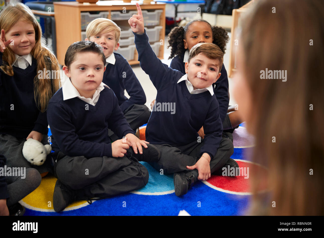 Blick über die Schulter, Lehrerin der Grundschule Kinder sitzen auf dem Boden im Klassenzimmer heben die Hände, um eine Frage zu beantworten Stockfoto