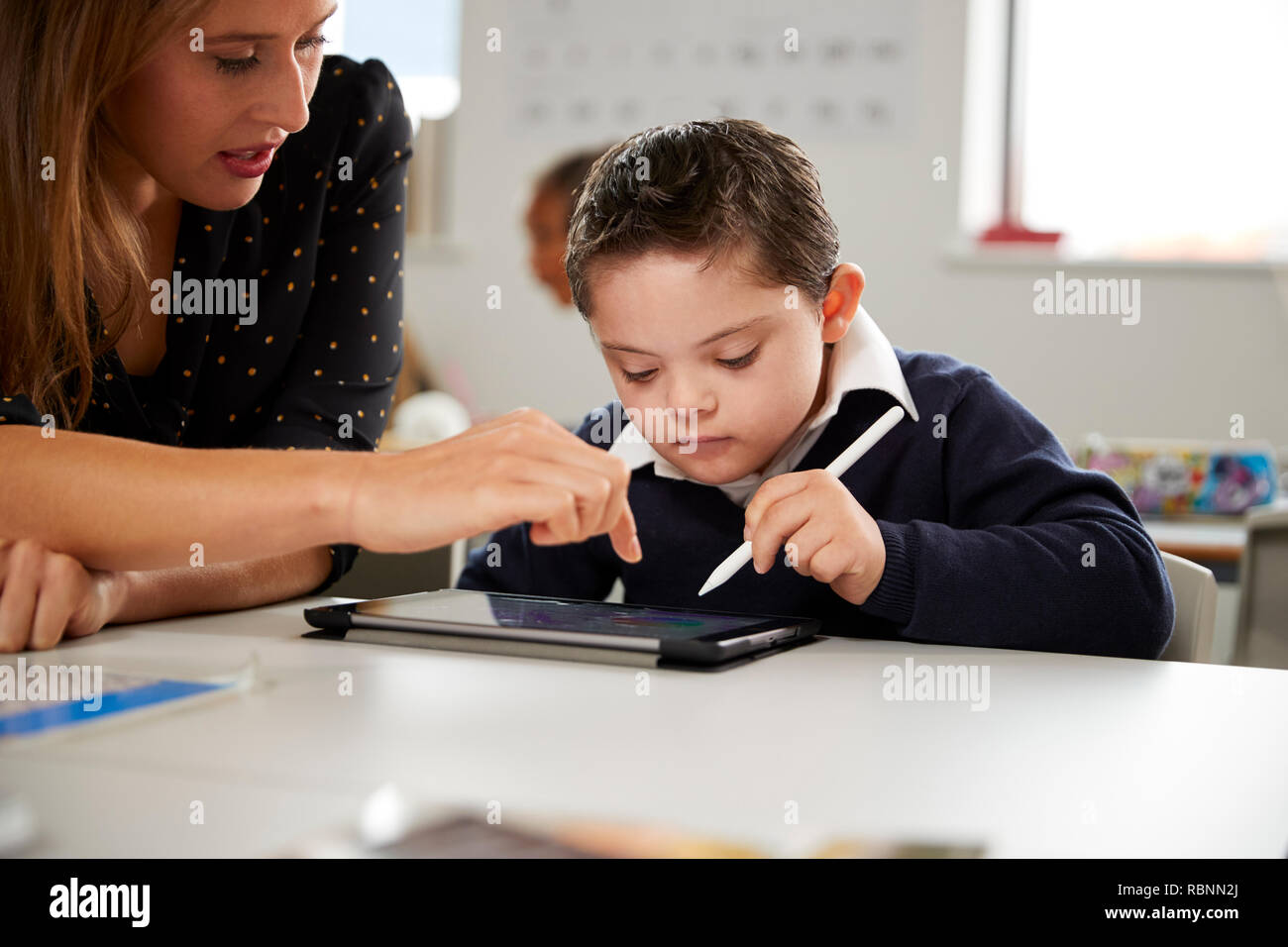 Junge weibliche Lehrer arbeiten mit einem Down Syndrom Schüler am Schreibtisch sitzend mit einem Tablet PC in einer Grundschule Klassenzimmer, Vorderansicht, in der Nähe Stockfoto