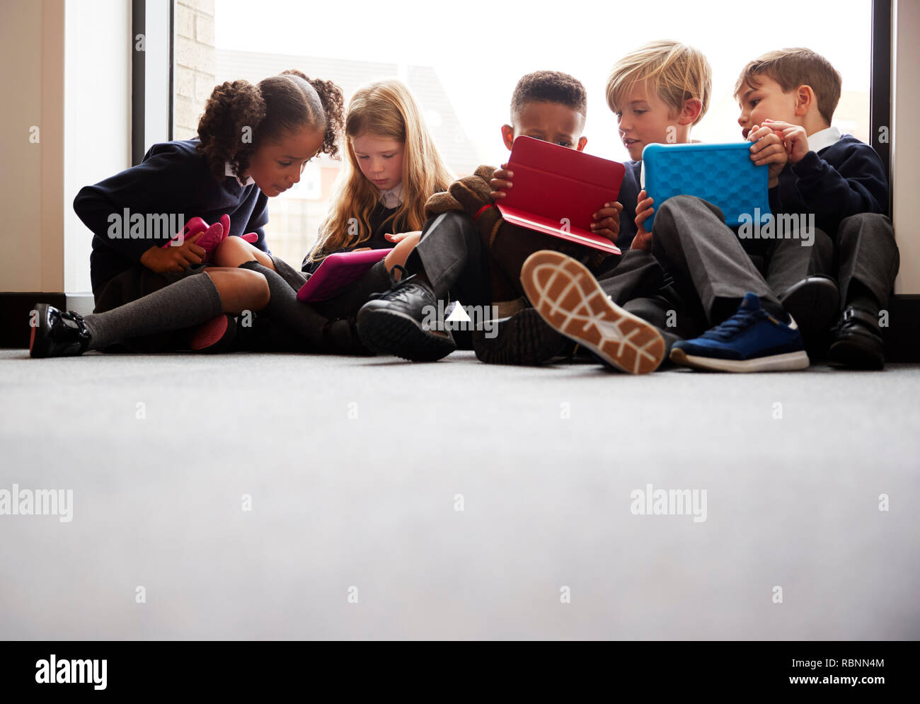 Low Angle View der Grundschule Freunde zusammen sitzen vor einem Fenster in einer Schule Flur am Tablet Computer suchen zusammen Stockfoto