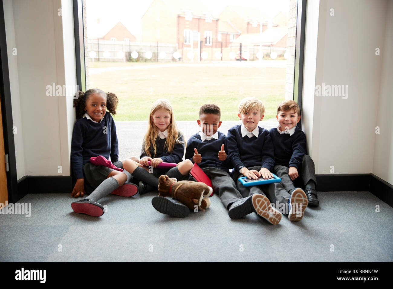 Fünf Grundschule Freunde mit Tablet-pcs auf dem Boden in einer Schule Korridor in den Pausen sitzen, Lächeln für die Kamera, selektiver Fokus Stockfoto