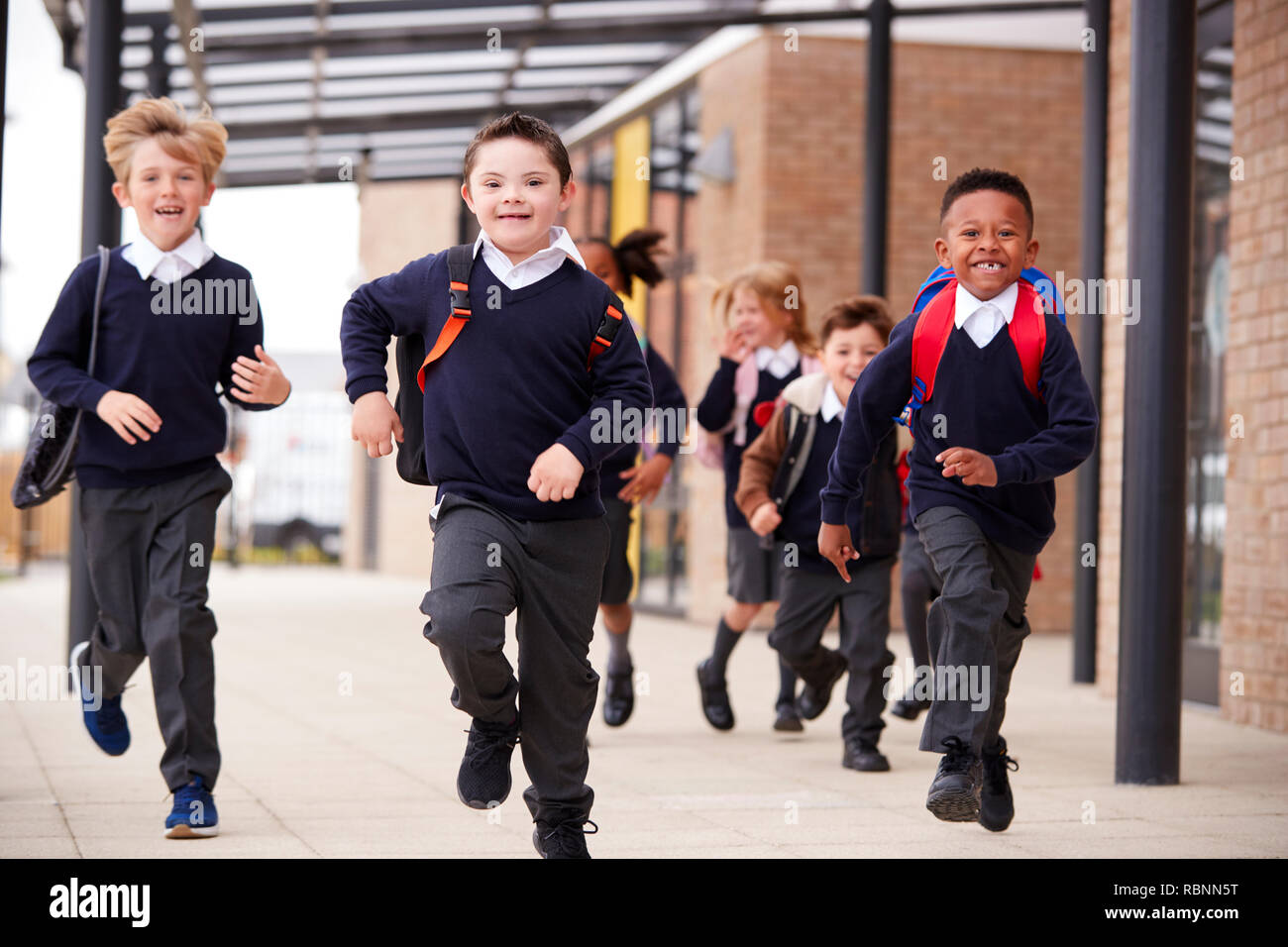 Aufgeregt Grundschule Kinder, das Tragen von Schuluniformen und Rucksäcke, die auf dem Gehweg außerhalb ihrer Schule Gebäude, Vorderansicht, in der Nähe Stockfoto