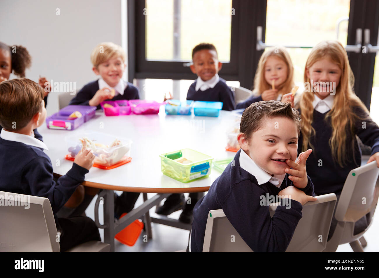 Ansicht der Grundschule Kinder sitzen gemeinsam an einem runden Tisch ihre Lunchpakete zu Essen, einige Drehen um die Kamera Stockfoto