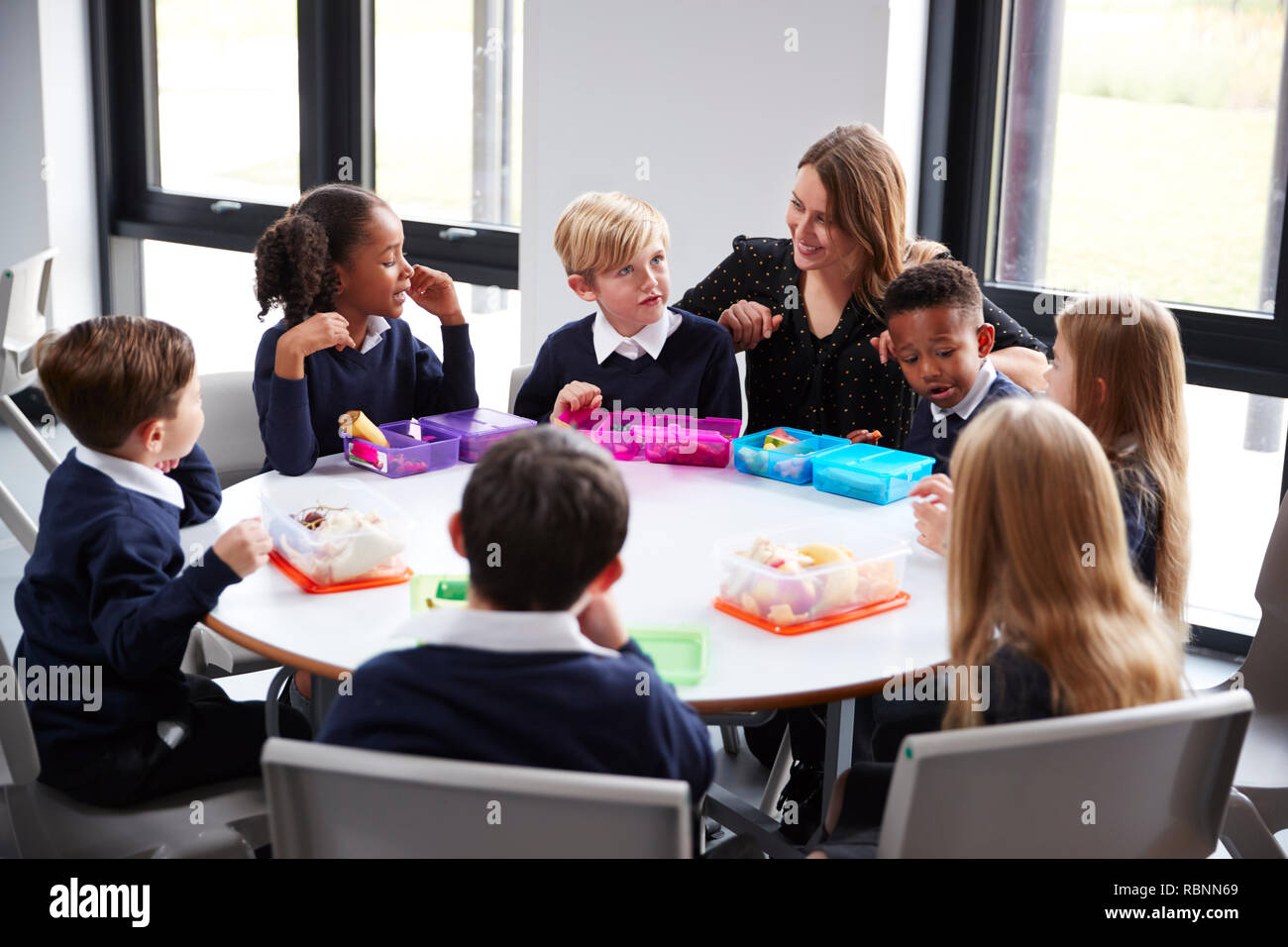 Lehrerin kniend zu einer Gruppe von Grundschule Kinder sitzen gemeinsam an einem runden Tisch essen ihre Lunchpakete zu sprechen Stockfoto