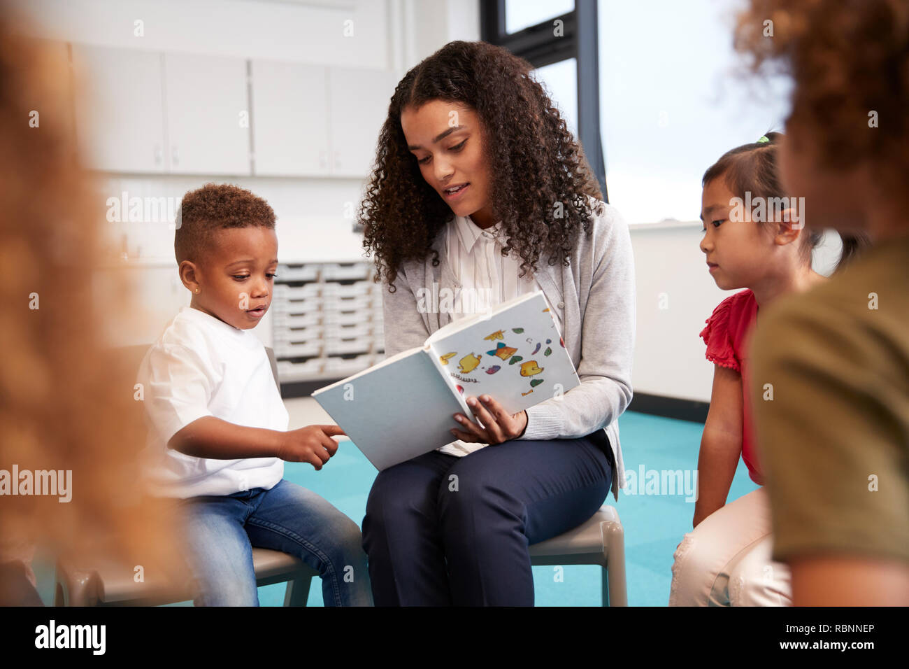 Infant School Junge in einem Buch von der Lehrerin gehalten, mit Kinder sitzen im Kreis auf Stühlen im Klassenzimmer, in der Nähe Stockfoto