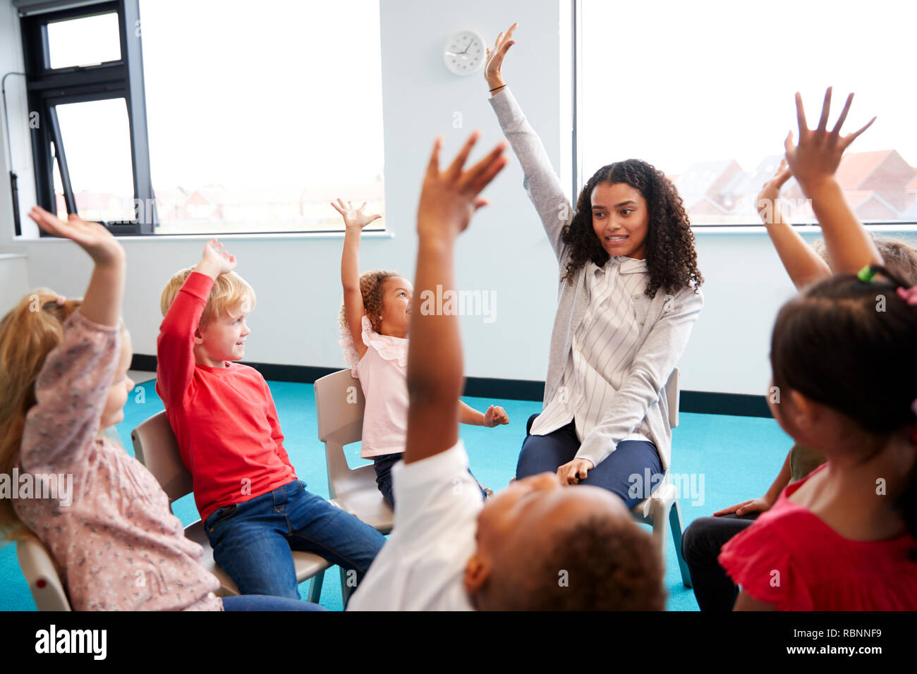 Eine Klasse für Kleinkinder Schulkinder sitzen auf Stühlen im Kreis im Klassenzimmer, heben die Hände mit ihrer Lehrerin, in der Nähe Stockfoto