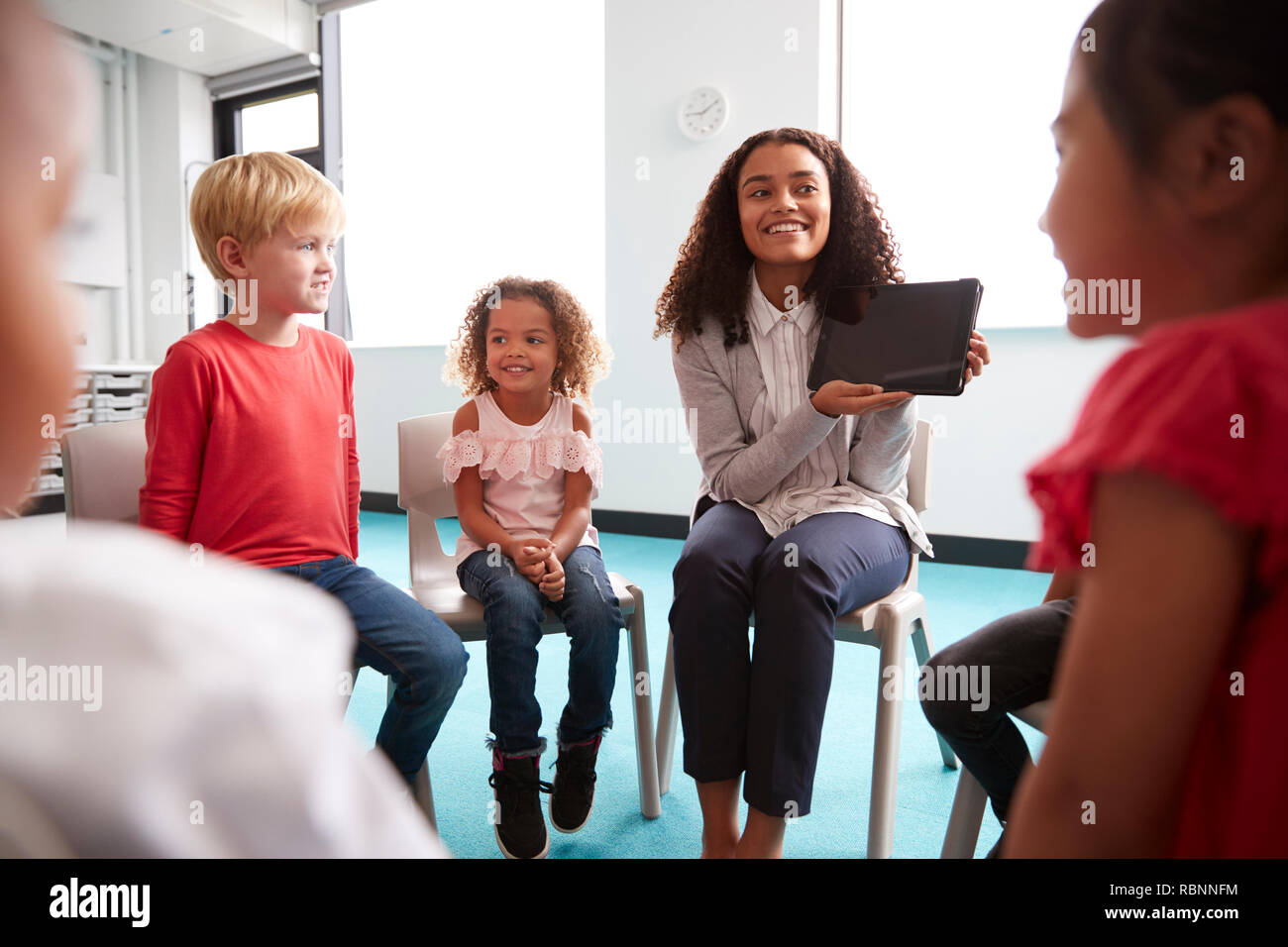 Blick über die Schulter, der lächelnden jungen weiblichen Lehrer zeigen einen Tablet-PC auf den neugeborenen Kindern, die im Kreis sitzen im Klassenzimmer, Blick über die Schulter Stockfoto
