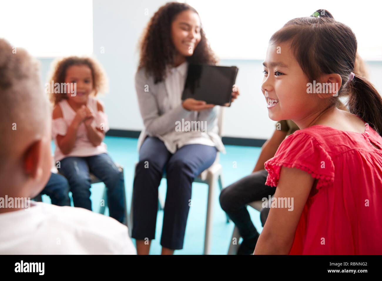 Blick über die Schulter, der lächelnden jungen weiblichen Lehrer zeigen einen Tablet-PC auf den neugeborenen Kindern, die im Kreis sitzen im Klassenzimmer, Blick über die Schulter Stockfoto