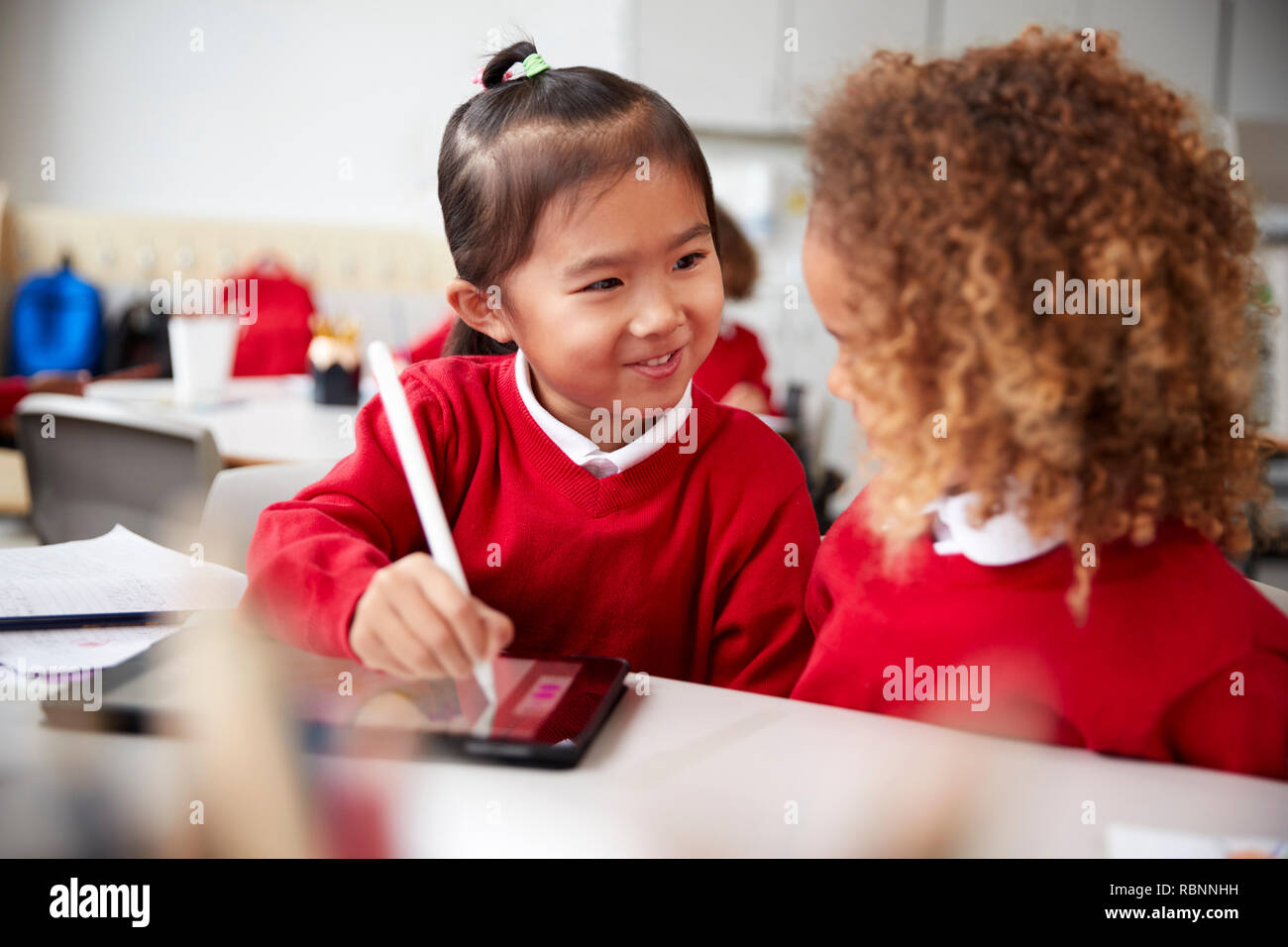 In der Nähe von zwei Kindergarten Schülerinnen das Tragen von Schuluniformen, sitzen an einem Schreibtisch in einem Klassenzimmer mit einem Tablet Computer und Stift, an jedem anderen suchen Lächeln Stockfoto