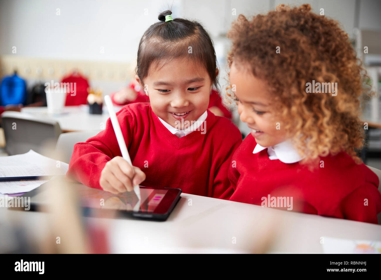 In der Nähe von zwei Kindergarten Schülerinnen das Tragen von Schuluniformen, sitzen an einem Schreibtisch in einem Klassenzimmer mit einem Tablet Computer und Stift, auf den Bildschirm zu schauen und lächeln Stockfoto