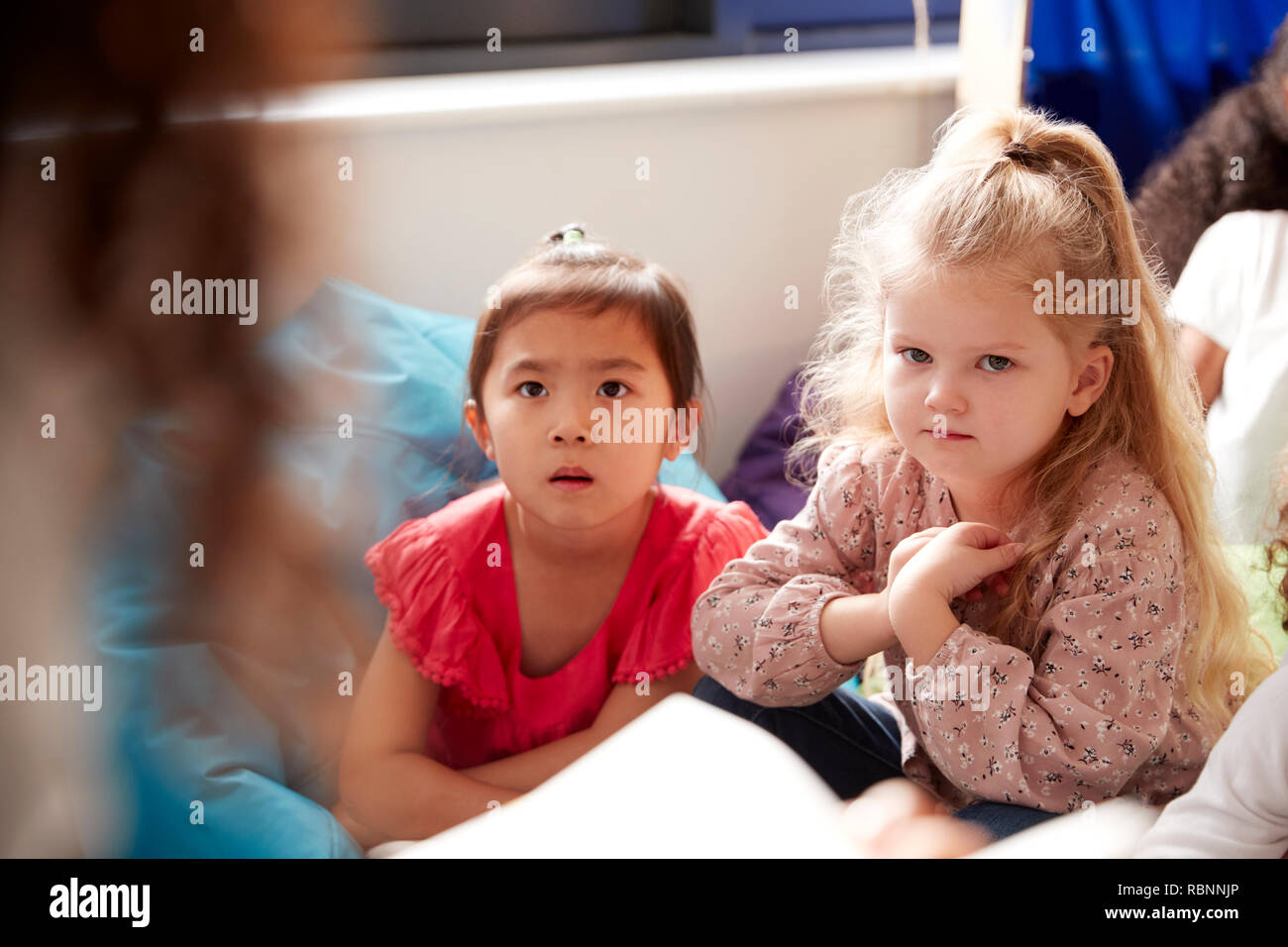 Zwei Infant School Mädchen sitzen auf Bohne Beutel in einer gemütlichen Ecke des Klassenzimmers hören auf ihre Lehrer eine Geschichte liest, Blick über die Schulter des Lehrers Stockfoto