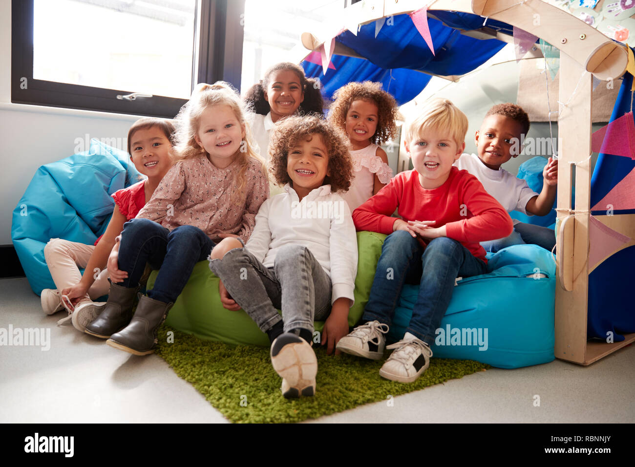 Eine multi-ethnische Gruppe der Kleinkinder Schulkinder sitzen auf Bohne Beutel in einer gemütlichen Ecke des Klassenzimmers, lächelnd auf Kamera, niedrigen Winkel, in der Nähe Stockfoto