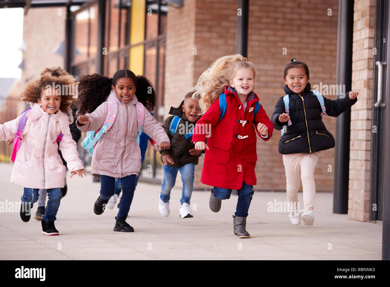 Glückliche junge Schule Mädchen, die Mäntel und tragen Schulmappen in einen Gehweg mit ihren Klassenkameraden außerhalb ihres Säuglings Schule Gebäude läuft Stockfoto