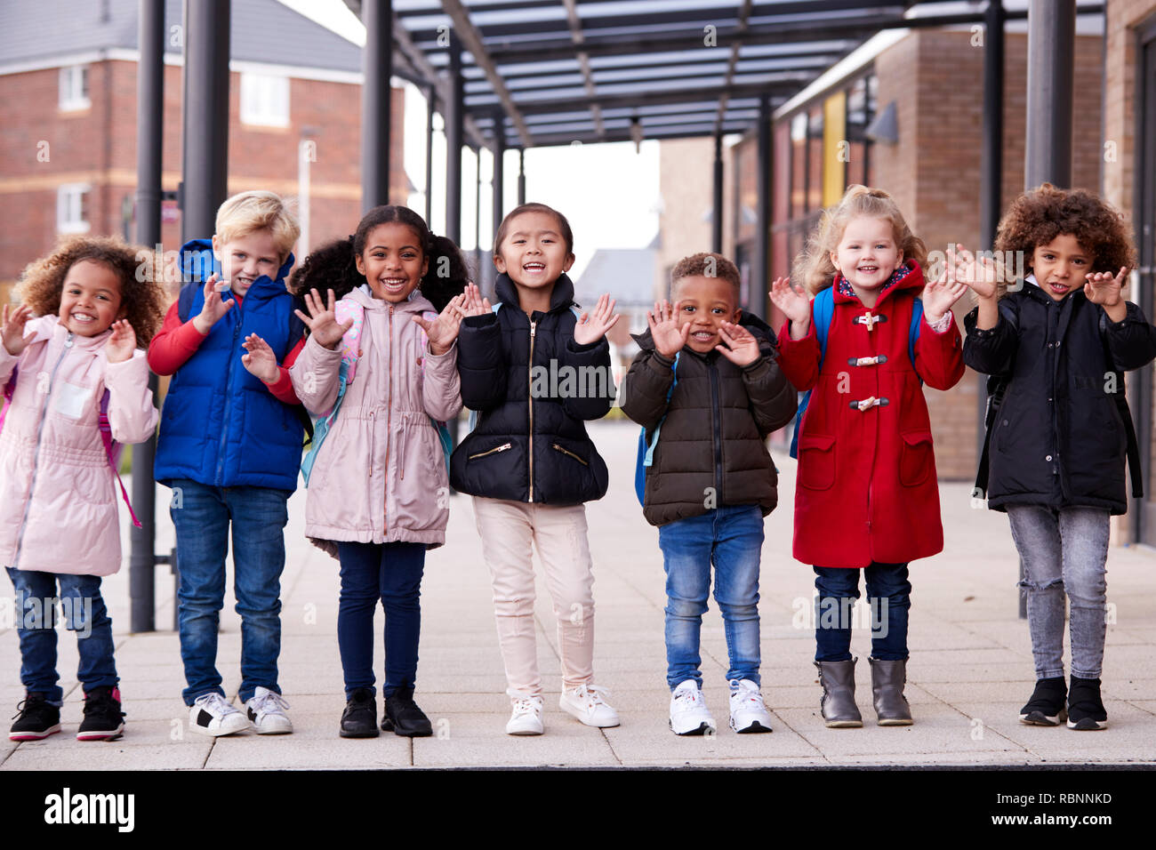 Eine Gruppe von lächelnden Jungen multiethnischen Schule Kinder Mäntel tragen und tragen schulmappen in einer Reihe stehen in Gang außerhalb ihres Säuglings Schule winken in die Kamera, volle Länge, Vorderansicht Stockfoto