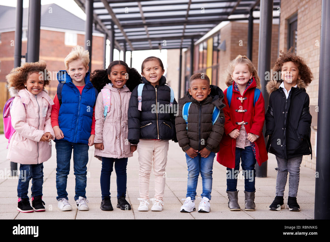 Eine Gruppe von lächelnden Jungen multiethnischen Schule Kinder Mäntel tragen und tragen schulmappen in einer Reihe stehen in Gang außerhalb ihres Säuglings Schule lächelnd in die Kamera, volle Länge, Vorderansicht Stockfoto