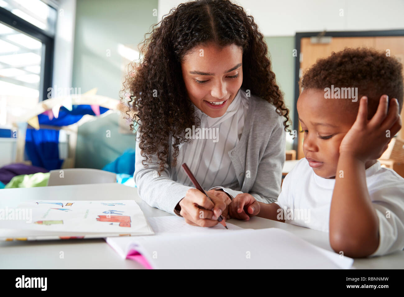 Weibliche Infant School Lehrer eine Arbeit an einem mit einem jungen Schüler, sitzen an einem Tisch mit ihm zu schreiben, in der Nähe Stockfoto