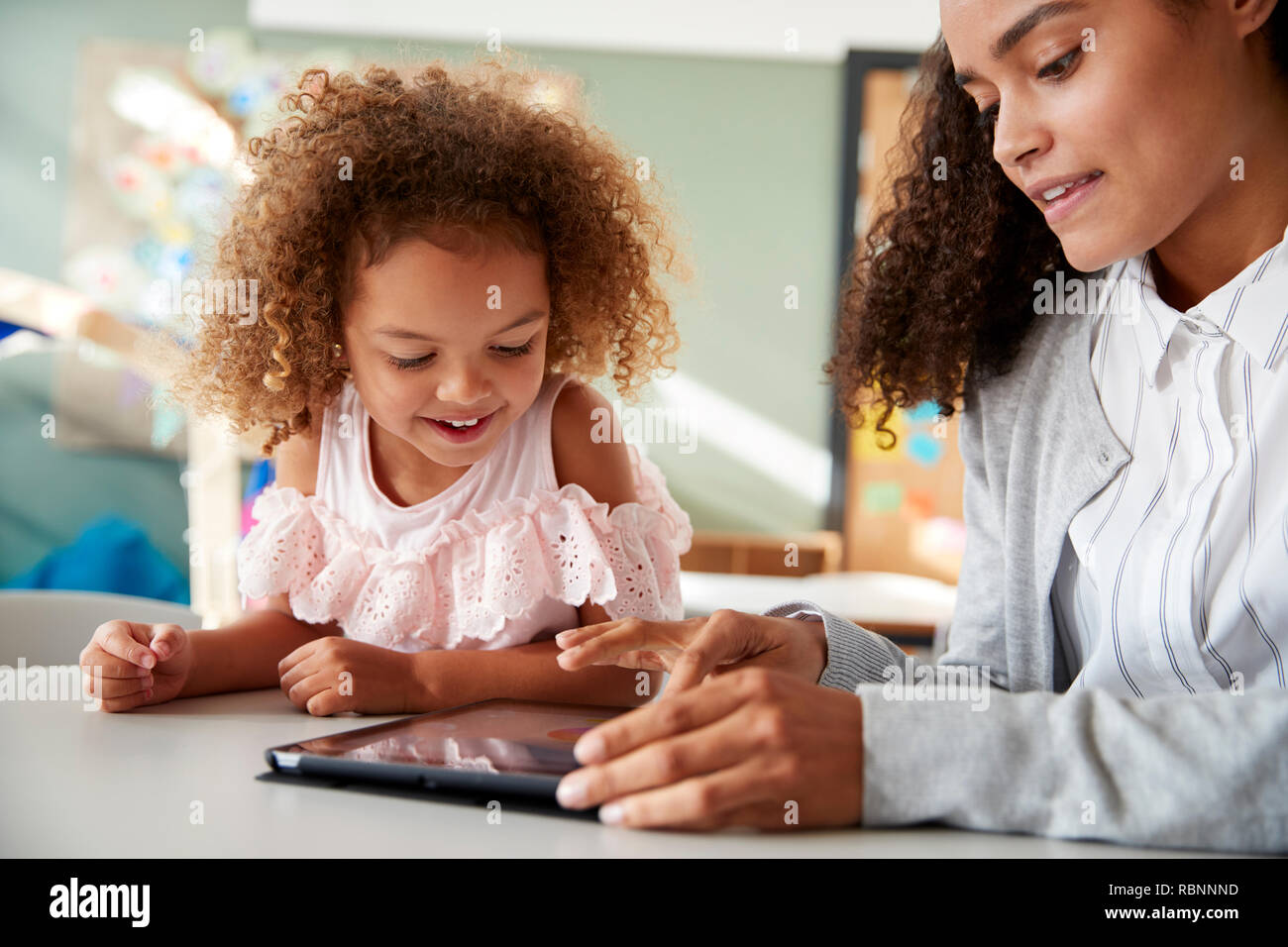 Weibliche Infant School Lehrer mit einem Tablet Computer arbeitet auf einer in einem Klassenzimmer mit einem Jungen gemischten Rennen Schulmädchen, selektiver Fokus, Nahaufnahme Stockfoto