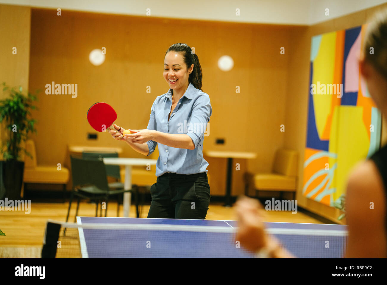 Eine attraktive asiatische Frau spielt Tischtennis mit einem Kollegen in einem stilvollen Coworking Space Office, wo Sie in einem Anlauf funktioniert. Sie lächelt. Stockfoto