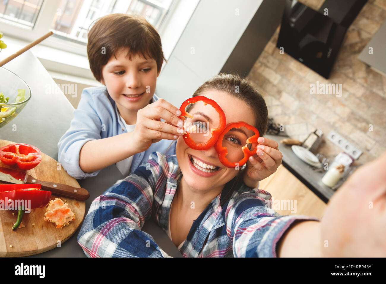 Mutter und Sohn zu Hause in der Küche zusammen selfie Fotos auf Handy boy Holding Paprika Kreise auf Mom's Augen Kamera lächeln Stockfoto