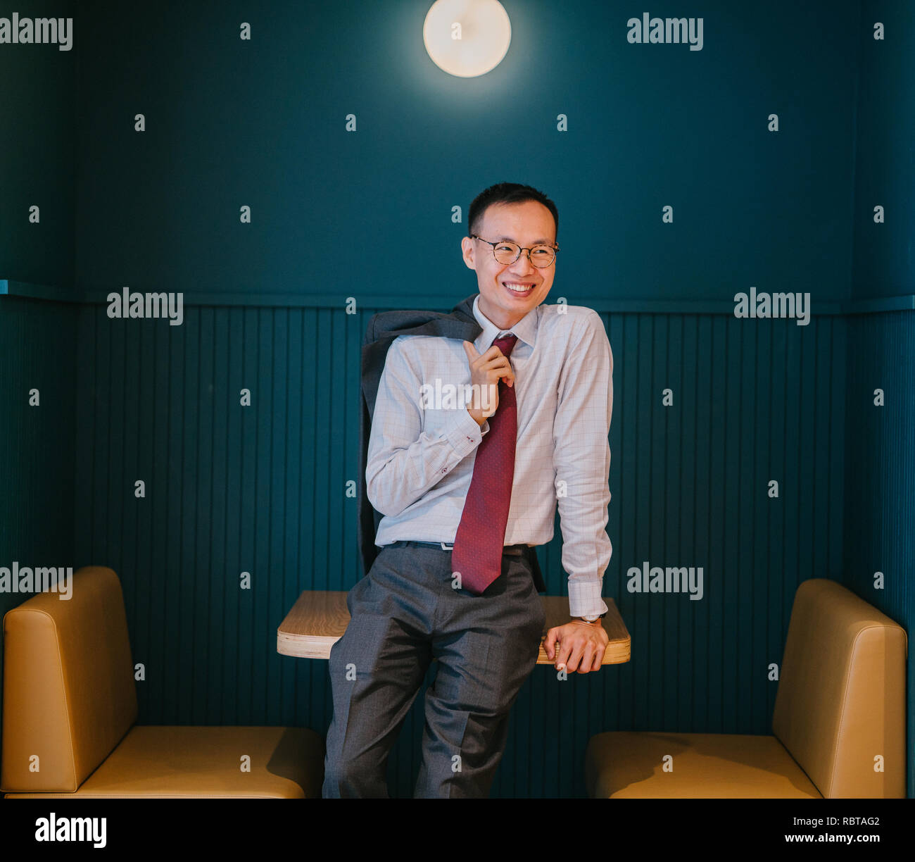 Porträt eines asiatischen Geschäftsmann in Anzug und Krawatte lehnte sich gegen ein trendiges blau stand in seinem Büro. Er ist selbstbewusst lächelnd. Stockfoto