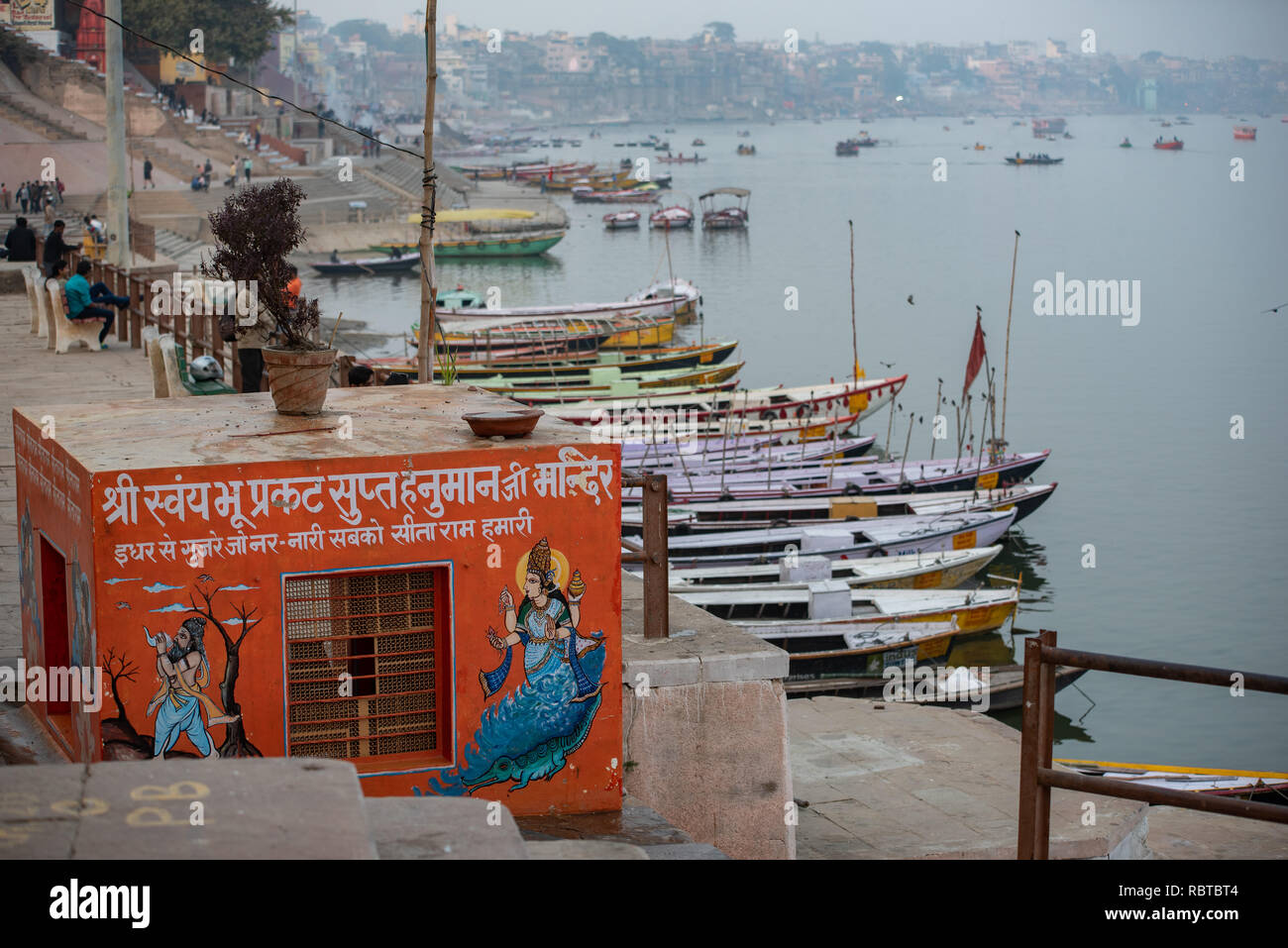 Allgemeine Ansicht des Ganges entlang der Ghats in Varanasi, Indien, mit einem kleinen orangefarbenen Tempel im Vordergrund und Boote im Hintergrund. Stockfoto