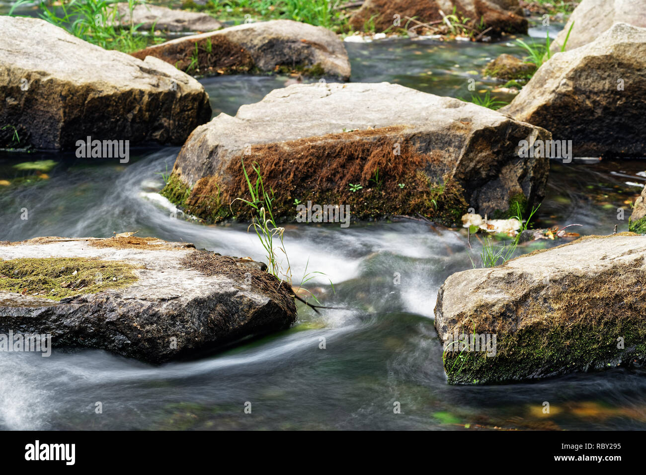 Detailansicht des fließenden Wasser eines kleinen Flusses, fließt das Wasser zwischen großen Steinen, teilweise mit Moos und Gras bewachsen, Langzeitbelichtung Stockfoto