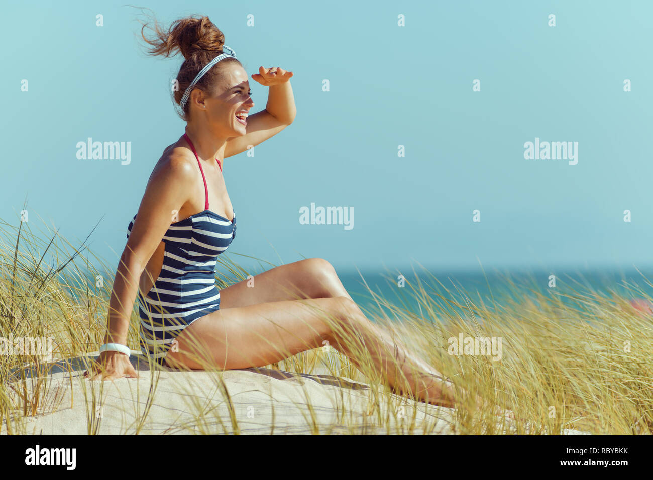 Gerne moderne Frau in gestreiften Badeanzug in die Ferne schauen, beim Sitzen am Meer. Ruhige Ferienhäuser Himmel blauer Himmel. Stockfoto Gerne moderne Frau in gestreiften Badeanzug in die Ferne schauen, beim Sitzen am Meer. Ruhige Ferienhäuser Himmel blauer Himmel. Stockfoto