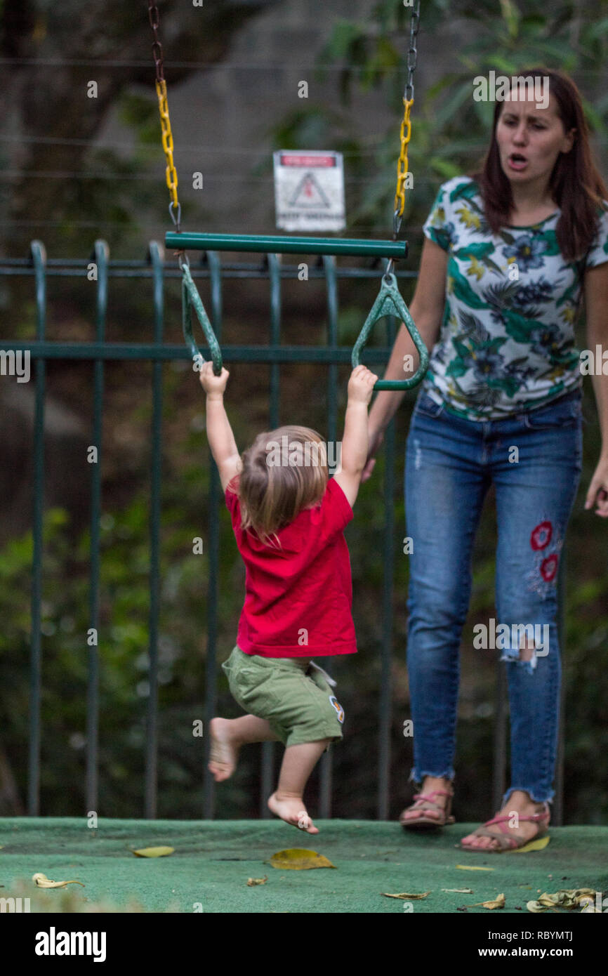 Ein Foto von einem starken toddler Training auf Pull up Fitnessstudio Ringe während seiner emotionalen Mutter jubelt Stockfoto