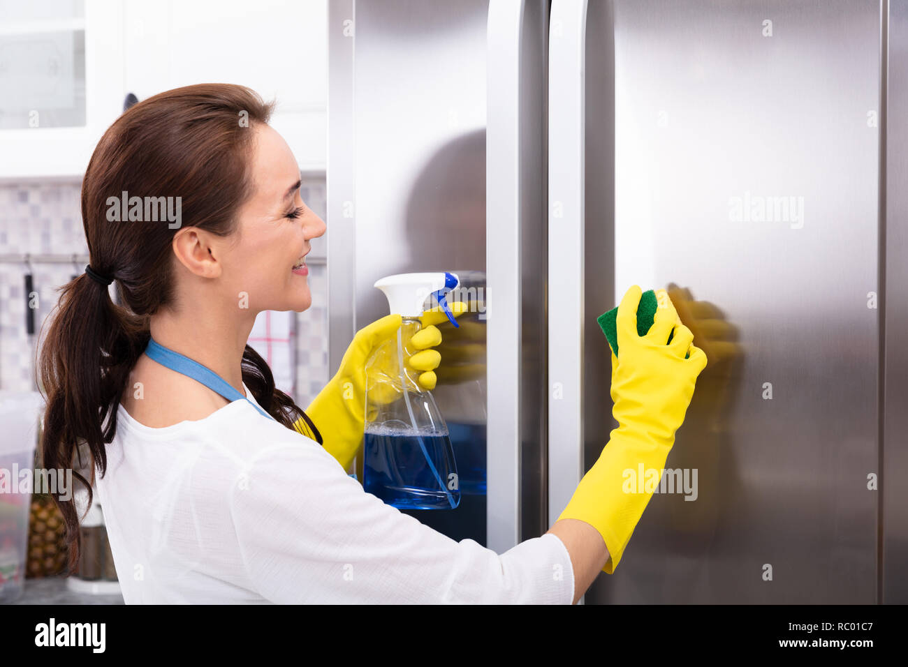 Glückliche junge Frau Reinigung Kühlschrank mit Schwamm und Flasche Spray Stockfoto