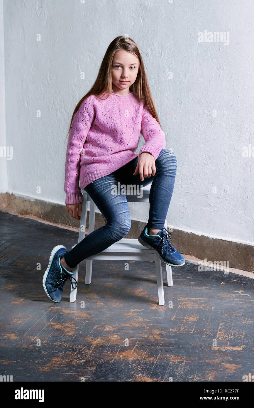 Zuversichtlich Kind mädchen mode Pose sitzt auf einem Stuhl, konkrete weißen Hintergrund. Elegantes Modell in rosa Pullover. Schöne glamourösen kid Teenager, lässig, u Stockfoto