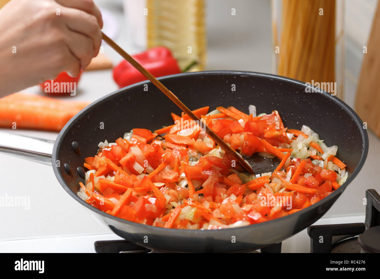 Hausmannskost. Eine Frau fries Zwiebeln, Möhren und Tomaten in einer heißen Pfanne mit Pflanzenöl. Close-up. Stockfoto