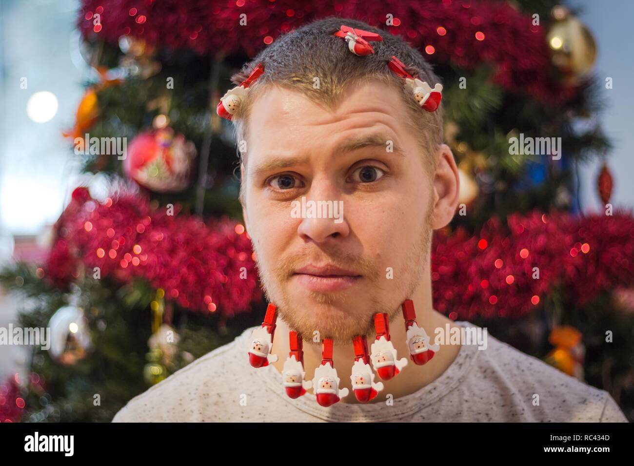 Ein junger und hübscher Kerl mit Bart auf dem Hintergrund der einen Weihnachtsbaum. Wäscheklammern von roter Farbe auf den Bart von der Kerl. Stockfoto