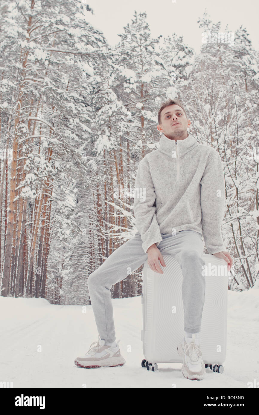 Porträt der jungen modische Mann in weißen Kleidern gehen auf Schnee holding Koffer und gehen weg Pinienwald. Winter Ferienhäuser Travel Concept. Outdoo Stockfoto