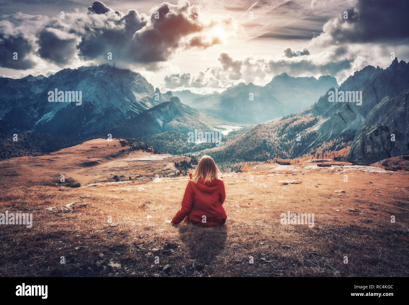 Junge Frau sitzt auf dem Feld gegen die majestätischen Berge bei Sonnenuntergang im Herbst in den Dolomiten, Italien. Landschaft mit Mädchen, bewölkter Himmel, orange Gr Stockfoto
