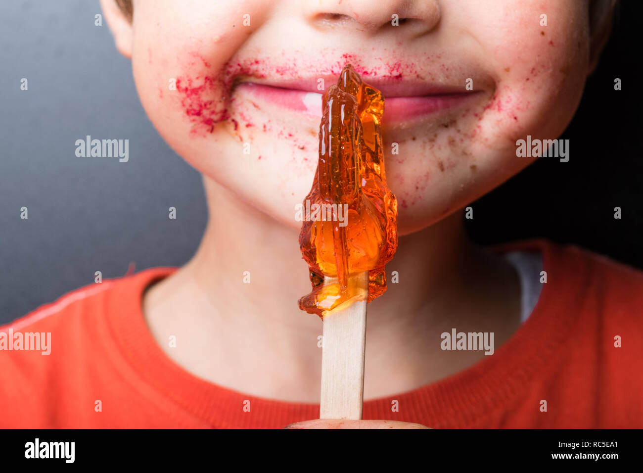 Boy saugt Lollipop, Baby isst - rot-orange Chupa Chups, Mund, rosa Lippen, Süßigkeiten in der Hand, das Gesicht halb Junge Stockfoto