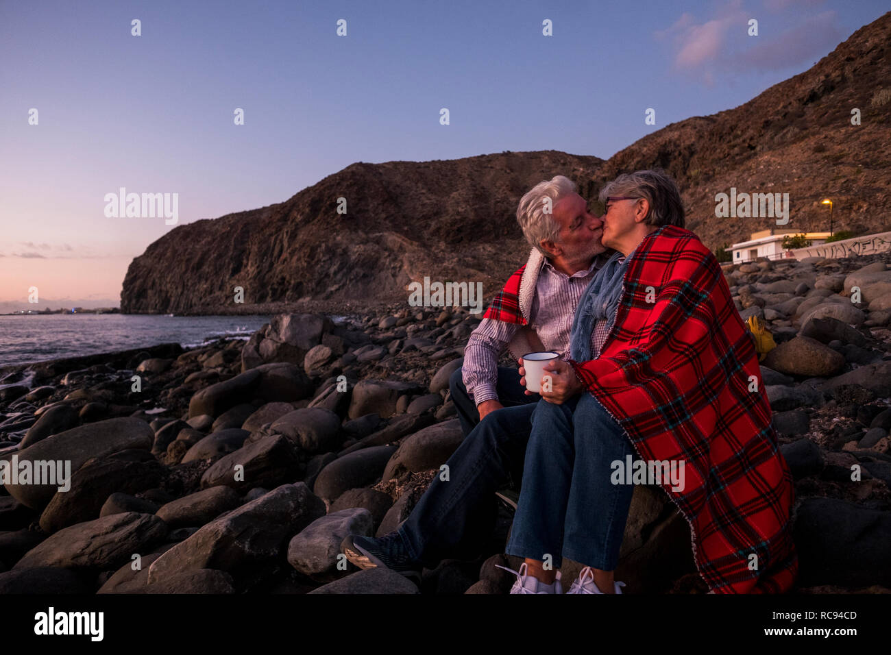 Ältere paar Küssen mit roter Decke auf Pebble Beach küssen bei Sonnenuntergang. Konzept für Urlaub, Freizeit, Liebe und für immer leben zusammen - Stockfoto