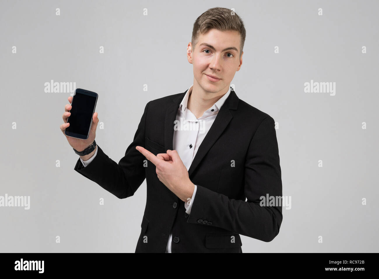 Portrait des jungen Mannes im schwarzen Anzug mit Finger auf Handy mit leeren Bildschirm auf weißem Hintergrund Stockfoto