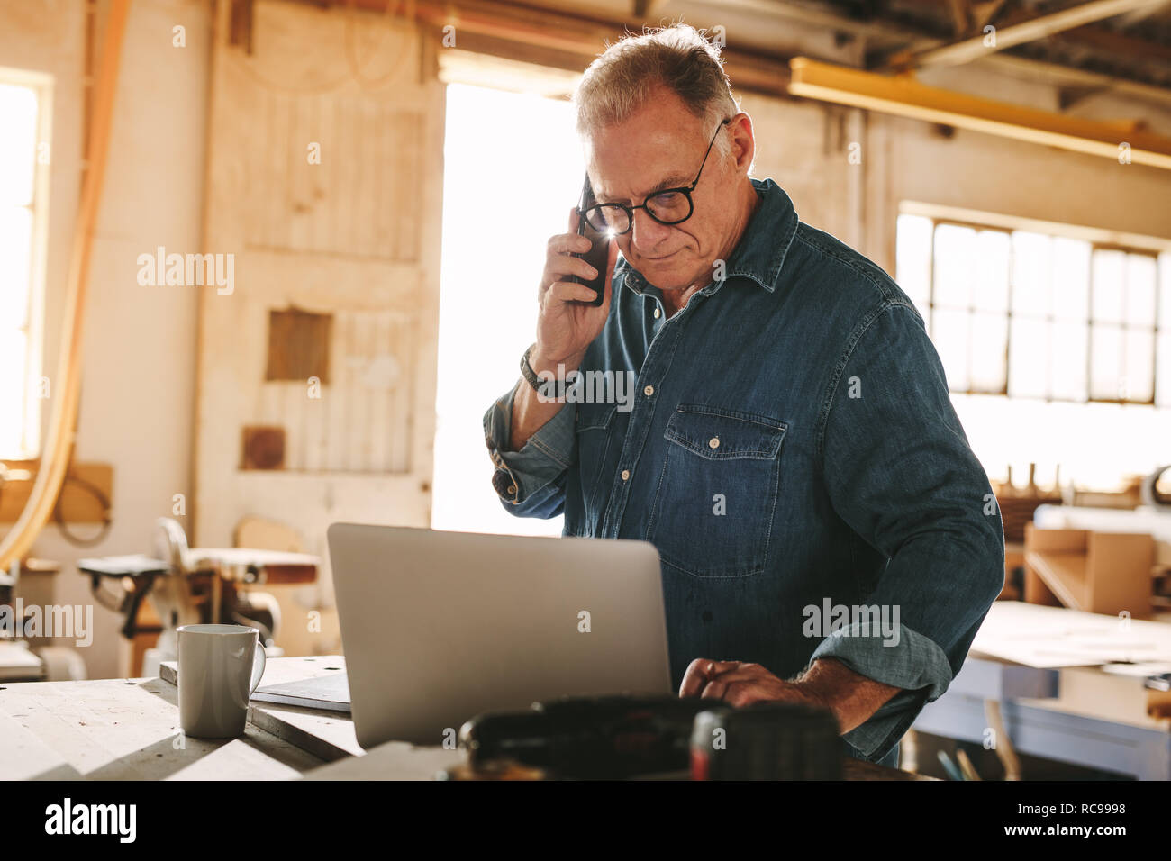 Ältere Menschen sprechen über Handy und Laptop auf dem Arbeitstisch. Reifen Tischler Arbeiten am Laptop und das Entgegennehmen von Anrufen in seiner Schreinerei Stockfoto