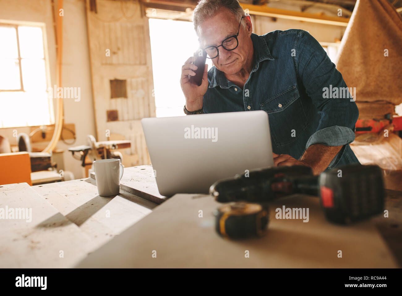 Älterer Mann mit Laptop und sprechen durch Smartphone in seiner Schreinerei. Im mittleren Alter Tischler Arbeiten am Laptop und das Entgegennehmen von Anrufen in seinem Stockfoto