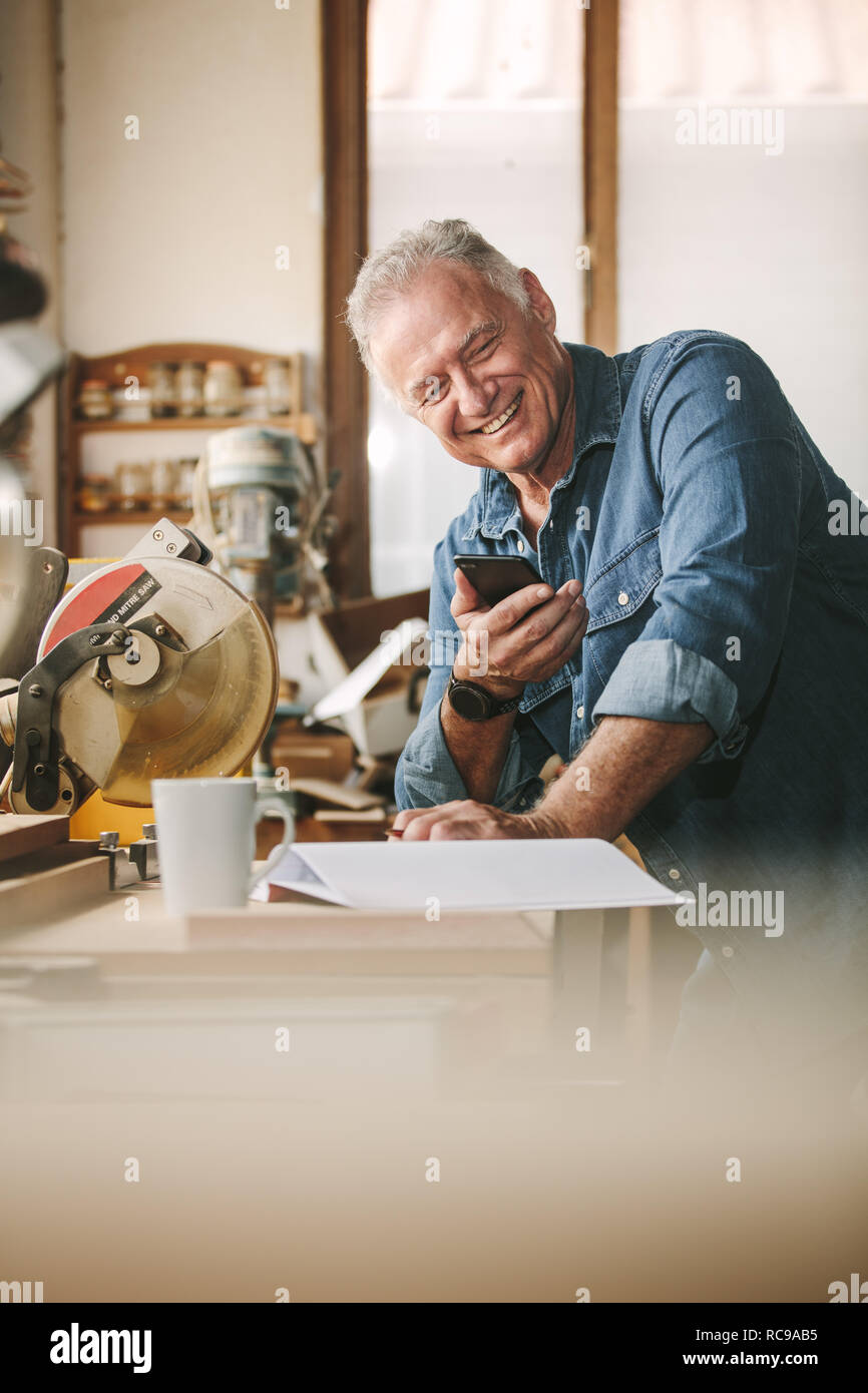 Senior Zimmermann in seiner Werkstatt mit Handy lächelnd. Reifer Mann in seiner Schreinerei lesen SMS-Nachricht auf sein Smartphone. Stockfoto