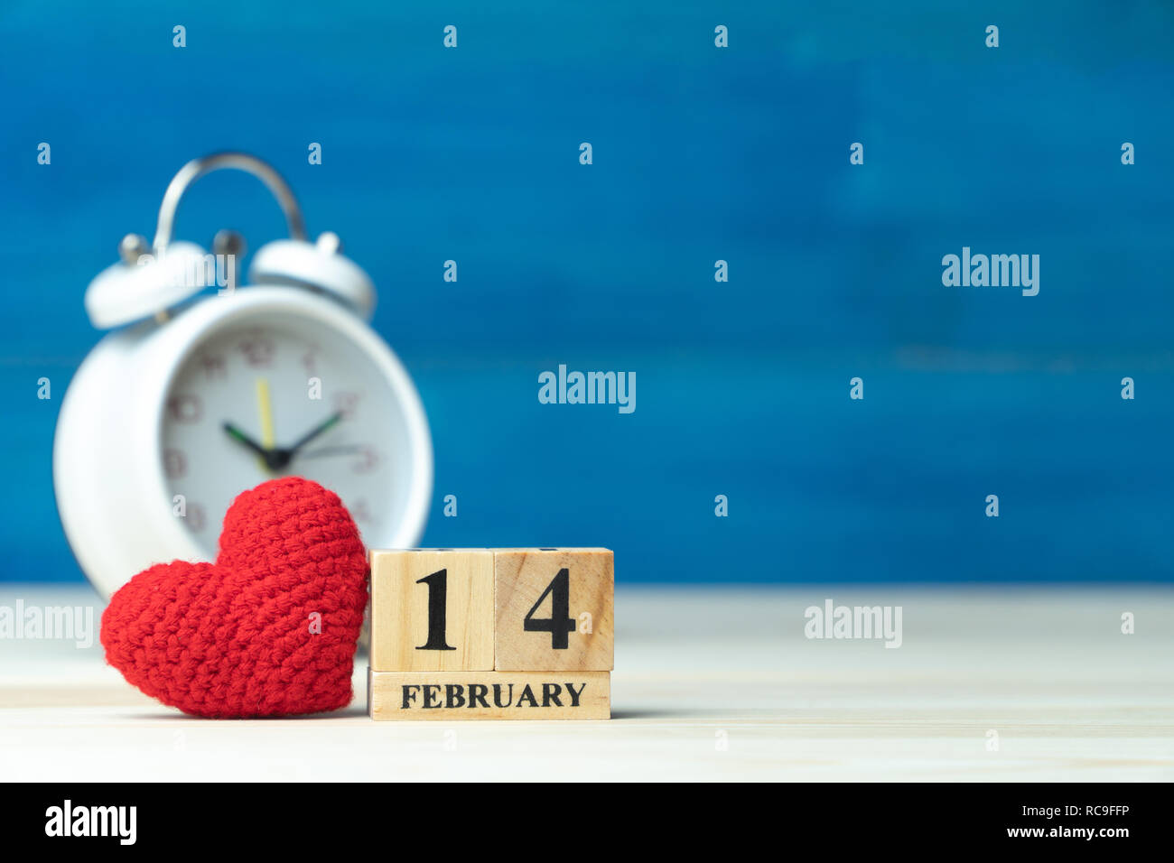 Valentines Tag Konzept. hand Garn machen Rotes Herz neben Holzblock Kalender auf Valentines Datum 14. Februar vor der weißen Wecker auf Holz gesetzt Stockfoto