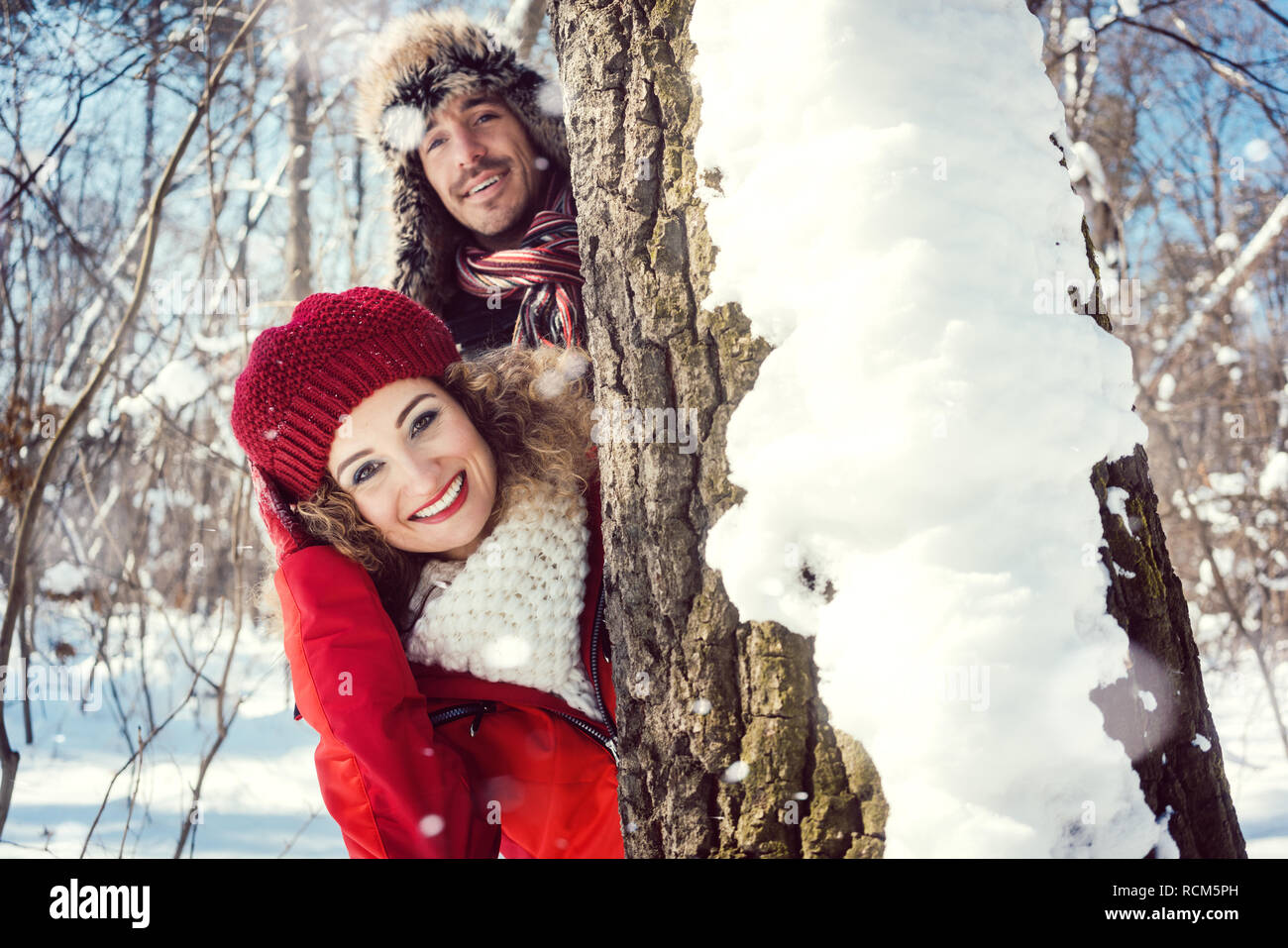 Verspieltes Paar im Schnee versteckt sich hinter einem Baumstamm Stockfoto