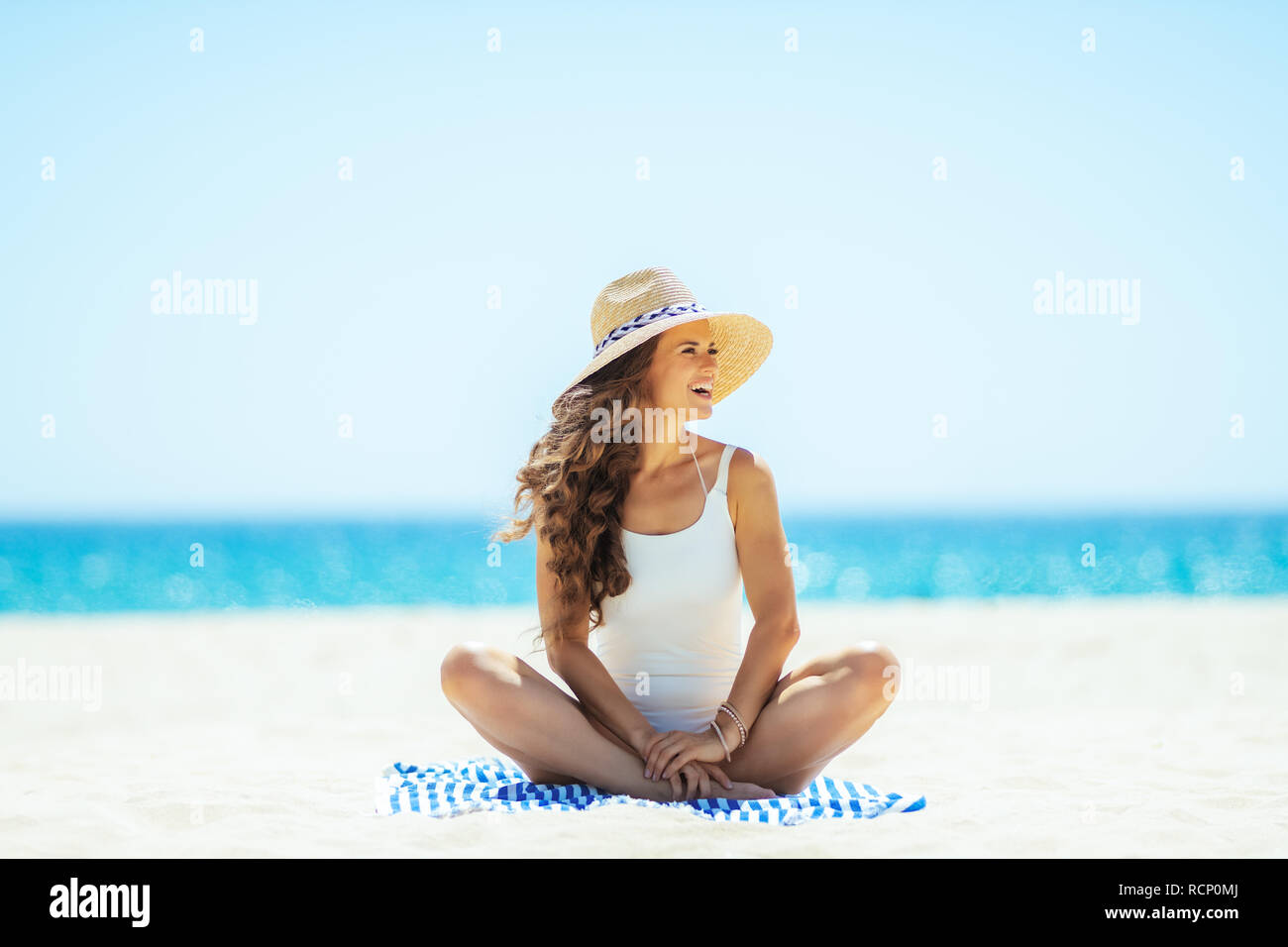 Gerne passende Frau in Weiß beachwear auf der Ocean Shore sitzen auf ein gestreiftes Handtuch und beiseite. Frau in Stroh Hut. Ruhige Ferienhäuser Himmel. Stockfoto Gerne passende Frau in Weiß beachwear auf der Ocean Shore sitzen auf ein gestreiftes Handtuch und beiseite. Frau in Stroh Hut. Ruhige Ferienhäuser Himmel. Stockfoto