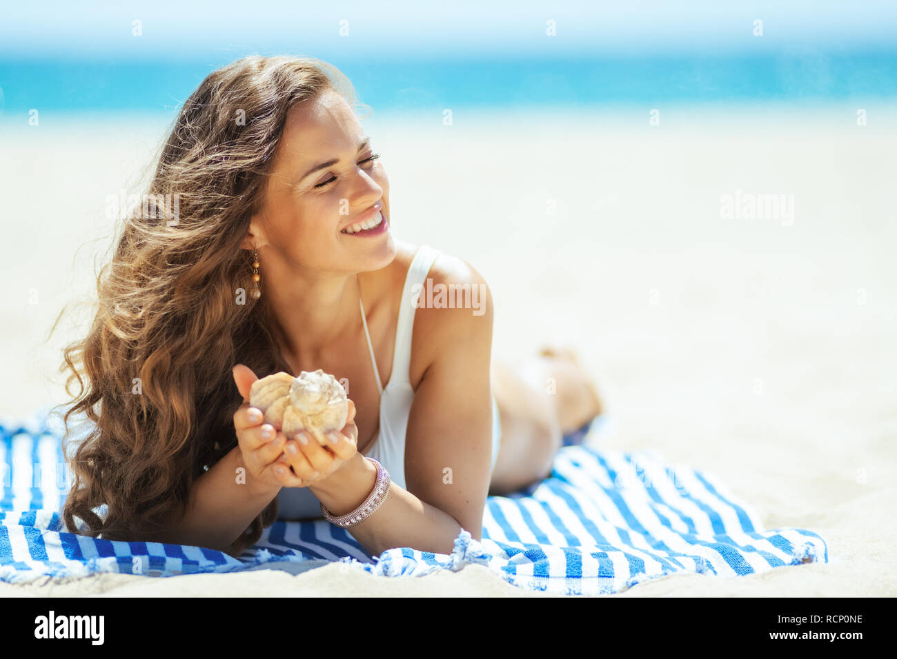 Entspannt gesunde Frau in weißen Badeanzug mit seashell Festlegung auf ein gestreiftes Handtuch auf der Seeküste. Erfolgreiche Beschießung im Urlaub am Strand. stresse Stockfoto Entspannt gesunde Frau in weißen Badeanzug mit seashell Festlegung auf ein gestreiftes Handtuch auf der Seeküste. Erfolgreiche Beschießung im Urlaub am Strand. stresse Stockfoto