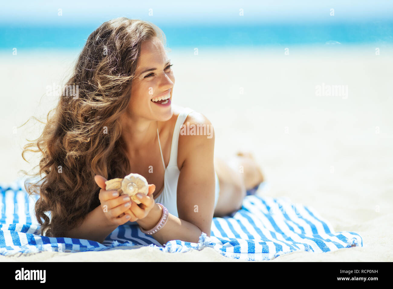 Glückliche gesunde Frau in weißen Badeanzug mit seashell Festlegung auf ein gestreiftes Handtuch und Blick auf die Küste. Erfolgreiche Beschießung im Urlaub an. Stockfoto Glückliche gesunde Frau in weißen Badeanzug mit seashell Festlegung auf ein gestreiftes Handtuch und Blick auf die Küste. Erfolgreiche Beschießung im Urlaub an. Stockfoto