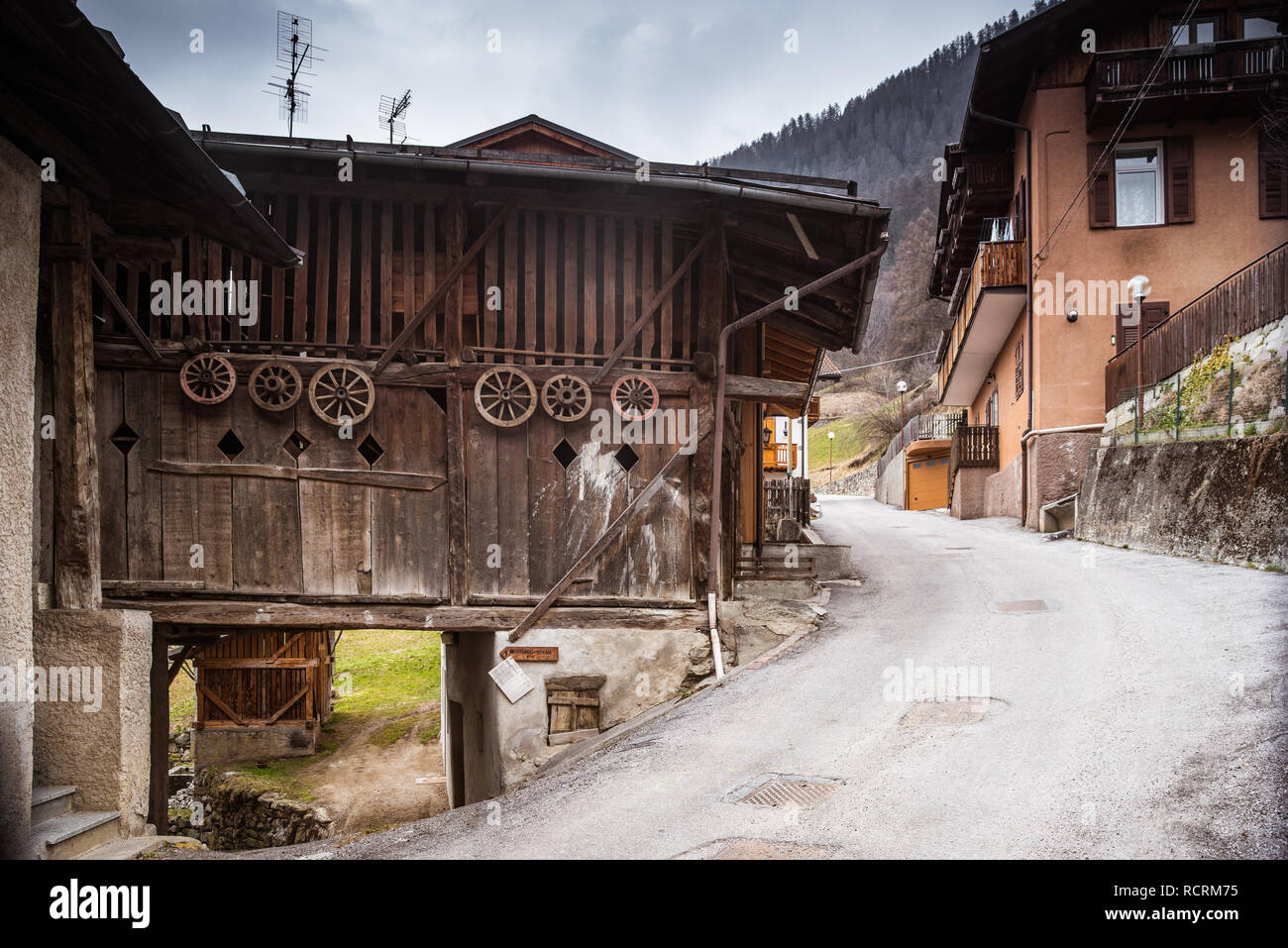 Alte hölzerne italienischen Häuser in einer kleinen Stadt Deggiano. Hölzerne Räder an der Wand des Gebäudes. Skigebiet Val di Sole, Brenta Dolomiten, Trento, Italien Stockfoto