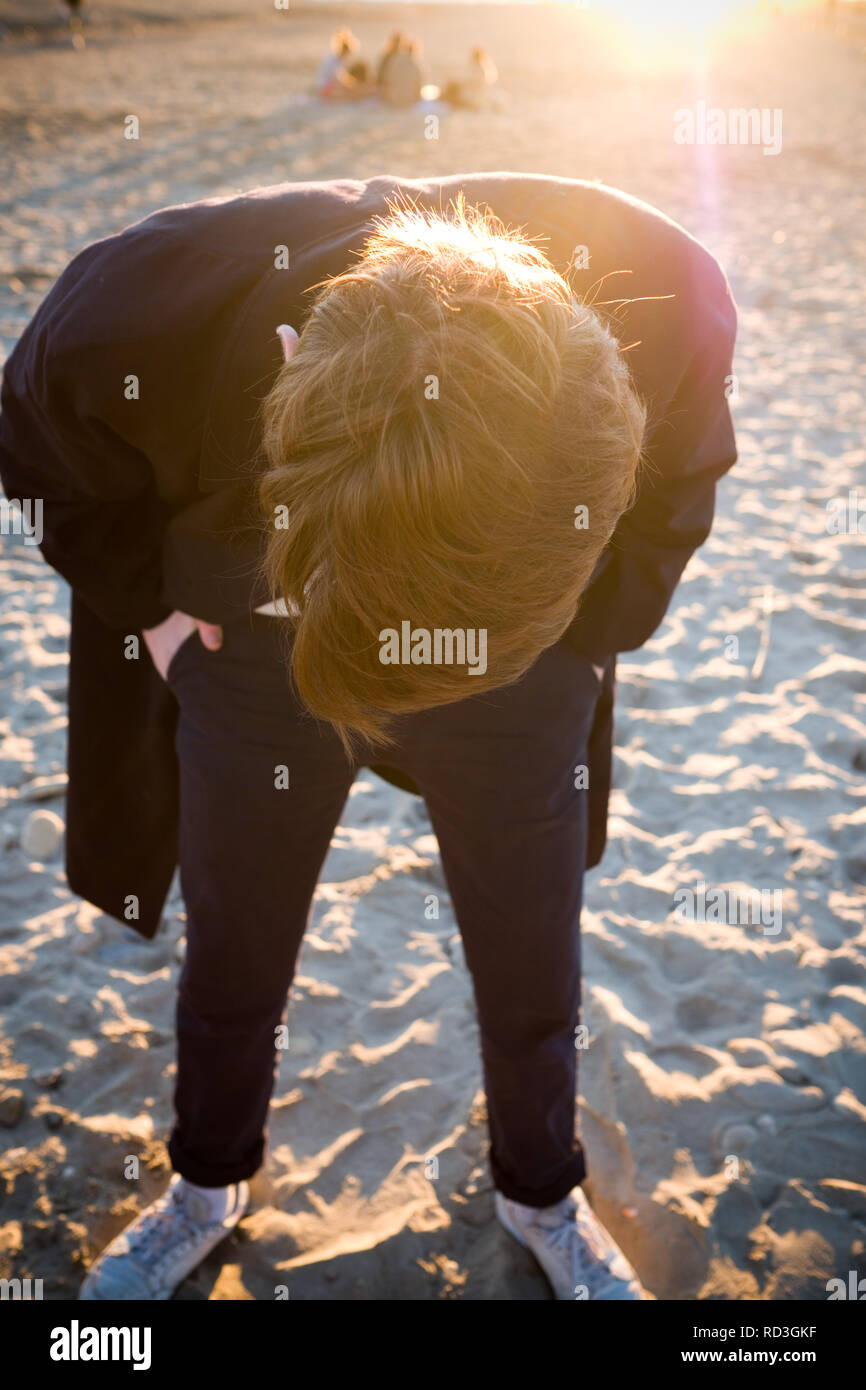 Ein männlicher Jugendlicher in einem schwarzen wollenen Mantel, bückt sich, während für etwas in den Sand am Strand bei einem Picknick auf der Suche Stockfoto