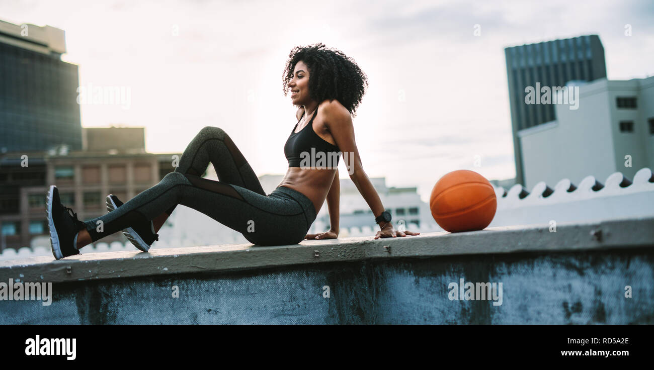 Frau in Fitness Tragen Sie sitzen entspannt auf der Dachterrasse Zaun mit einem Basketball neben ihr. Lächelnd weibliche Athleten Übungen in den Morgen auf der Terrasse Stockfoto