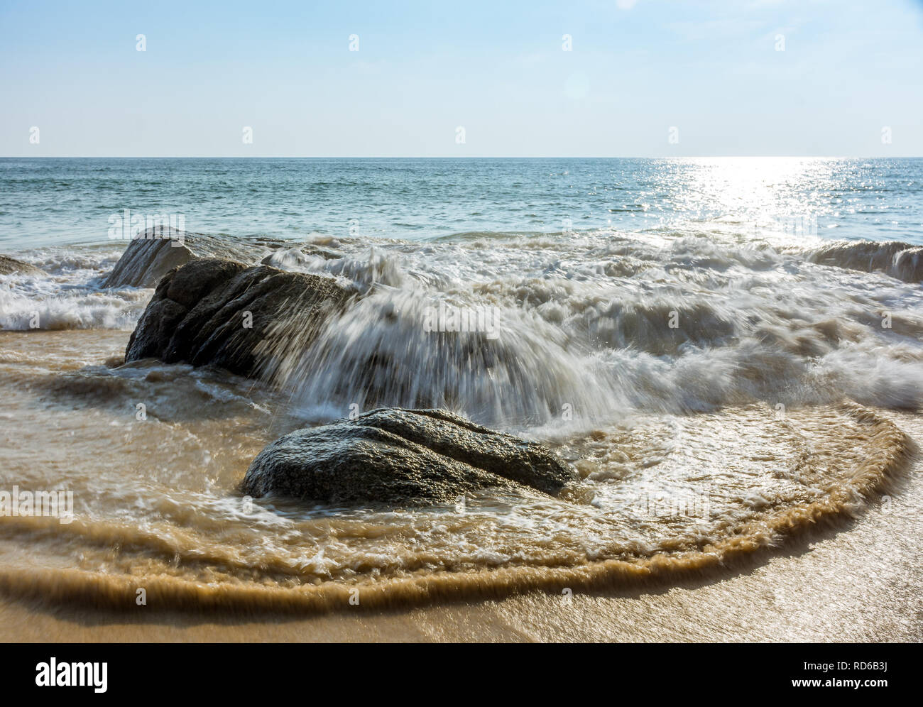Eine kleine Welle von einem ruhigen Meer stürzt über grosse Felsen an einem Sandstrand. Stockfoto
