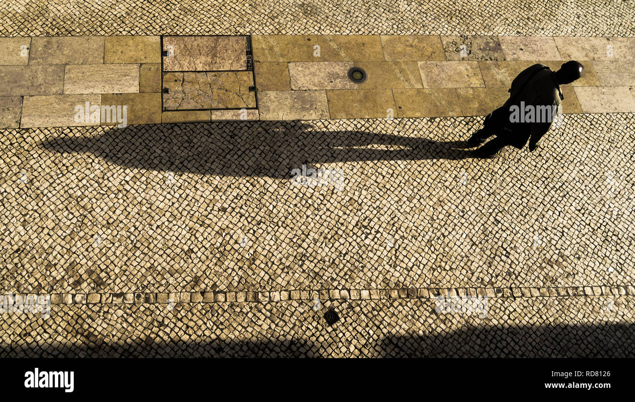 Lissabon, Portugal. Mann mit einem langen Schatten, der im Nachmittagslicht die Rua Barros Queirós nahe dem Rossio-Platz hinuntergeht. Typischer Kalksteinpflaster. Stockfoto